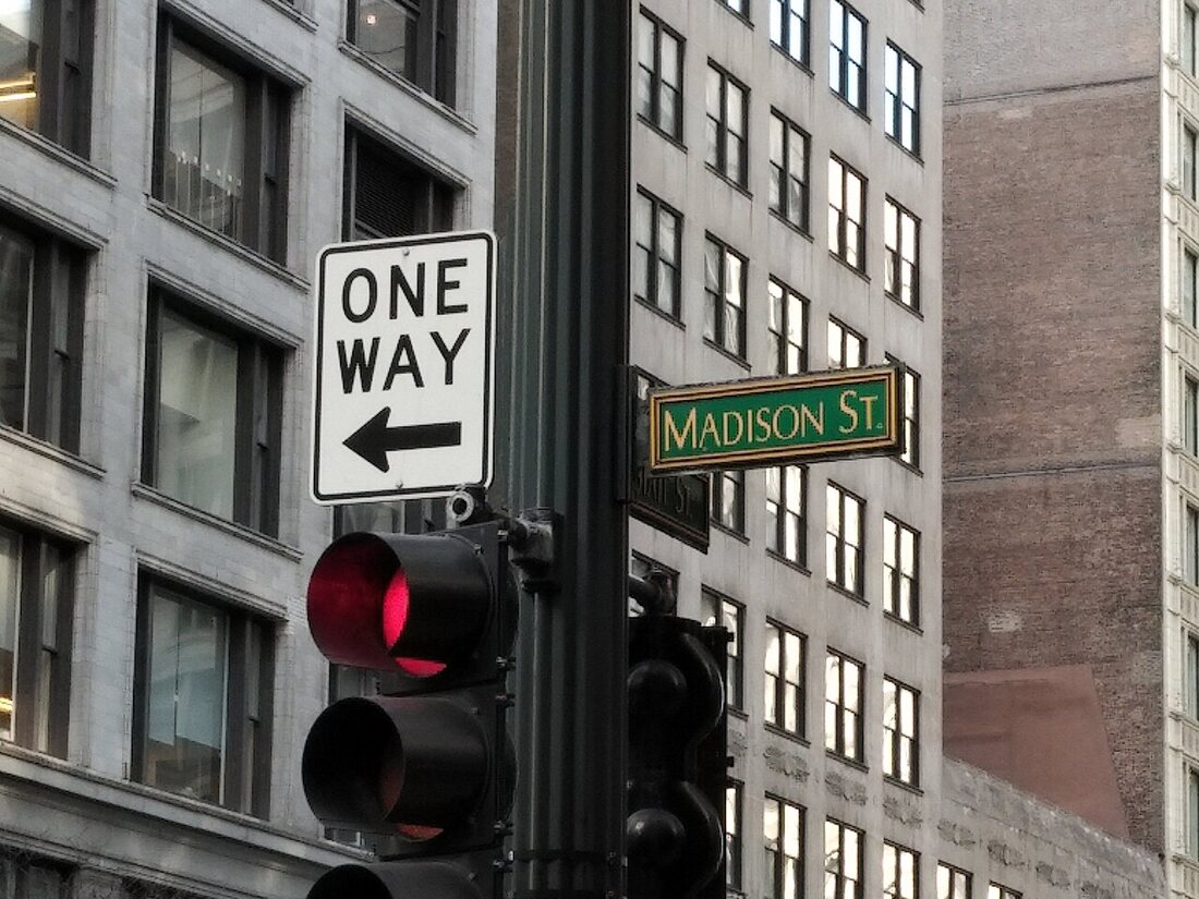 A black and white One Way sign with a left arrow mounted on a traffic light pole at Madison Street in Chicago, office buildings behind